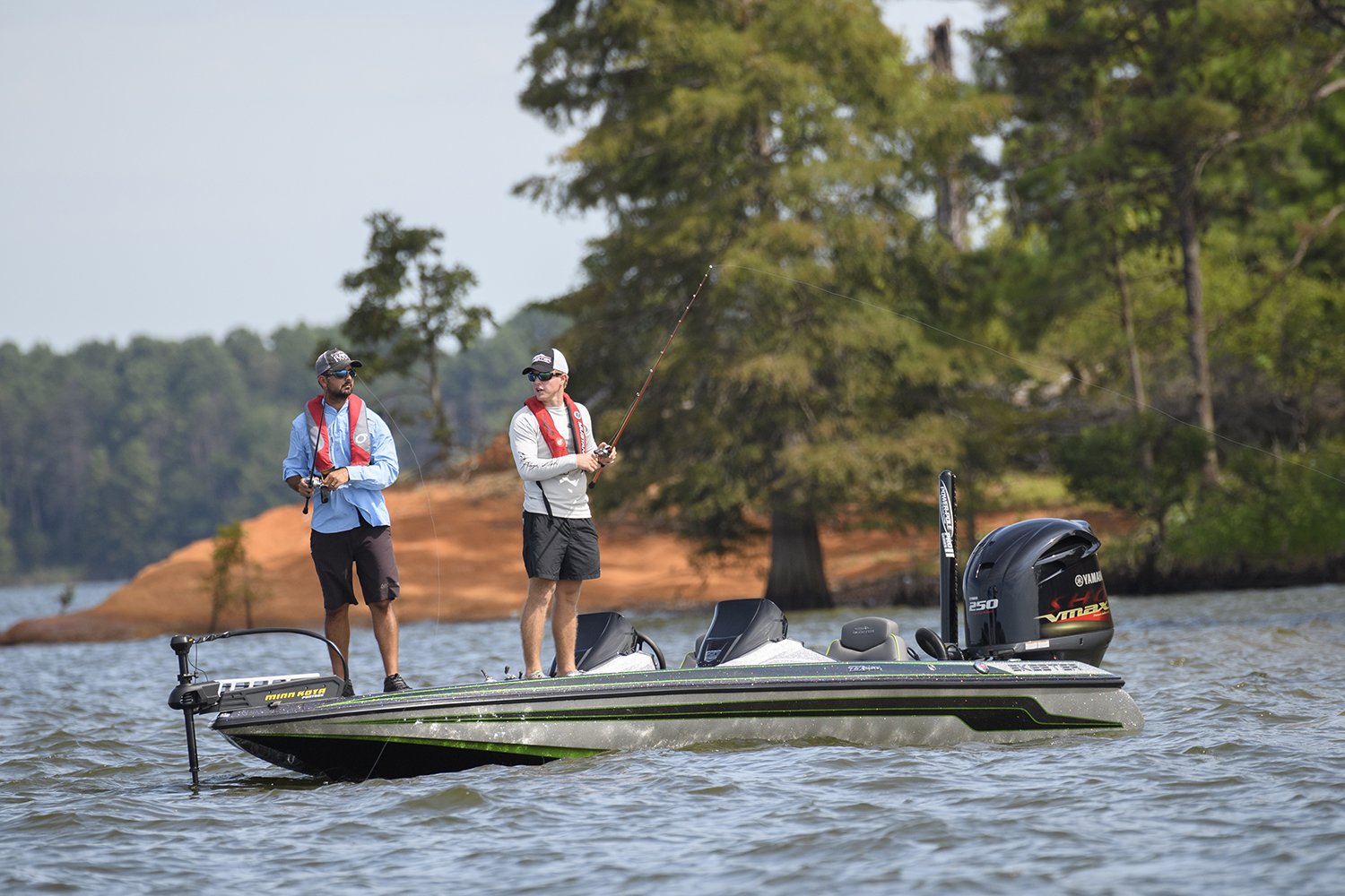 Two people wearing life vests are fishing from a boat on a lake, with trees and a sandy shoreline visible in the background.