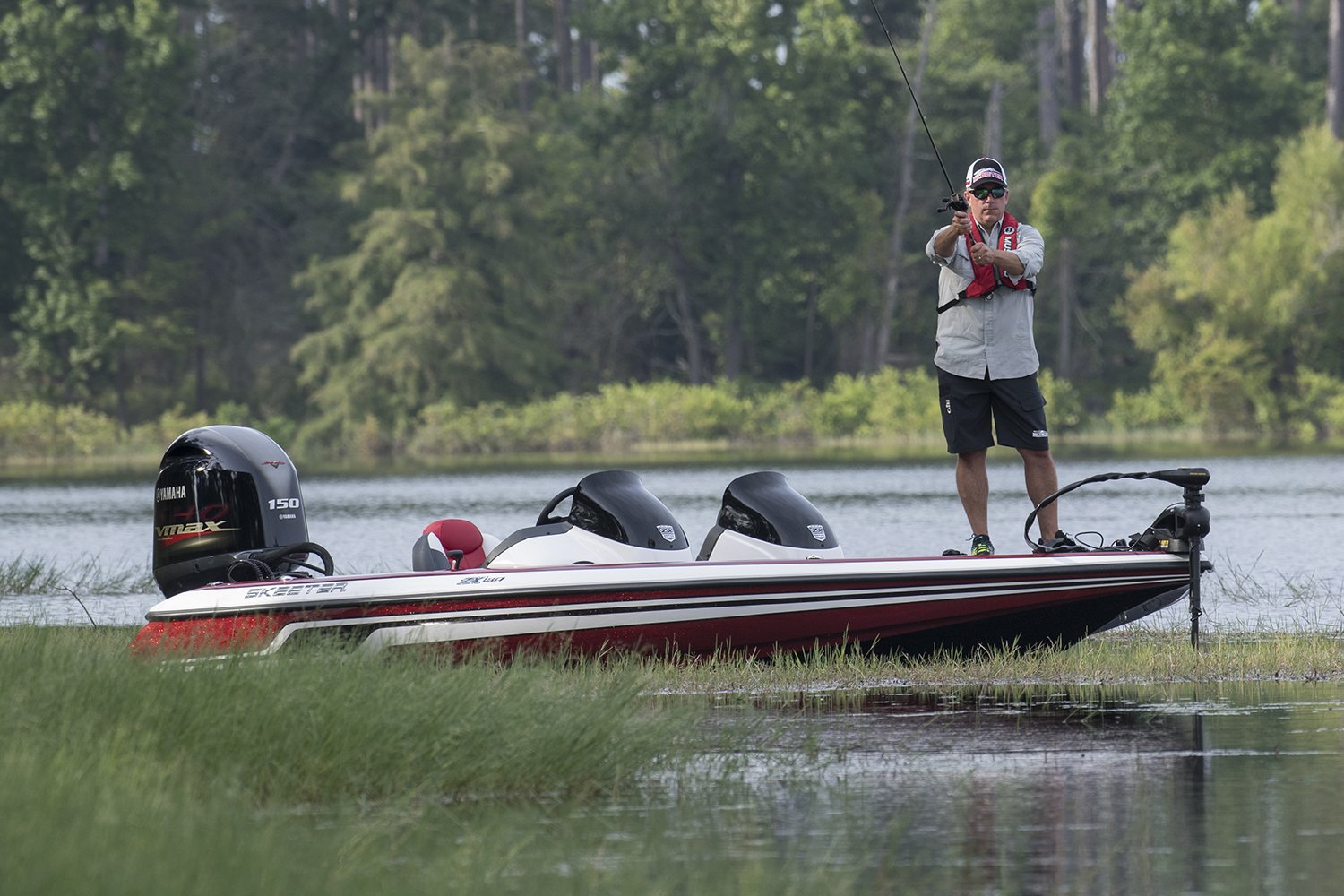 Person standing on a bass boat fishing with a rod on a calm lake, surrounded by grassy water and trees in the background.