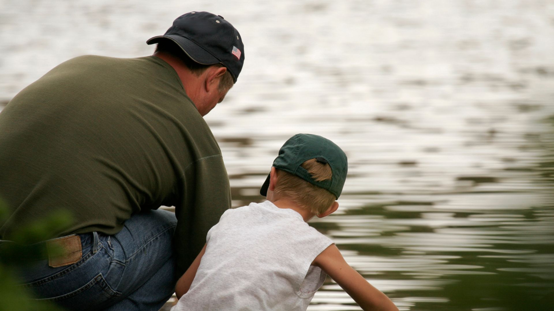 An adult and a child wearing caps crouch by the water's edge, looking at the lake.