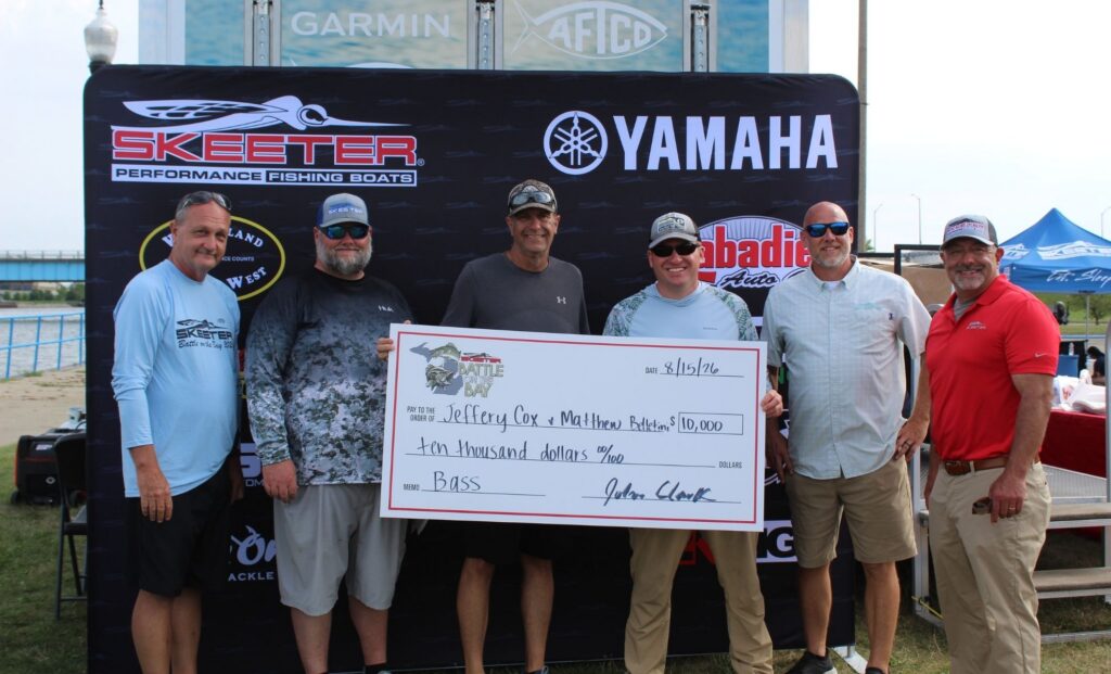 Six men stand outdoors holding an oversized check for $10,000 made out to Jeffrey Cox and Matthew Bates, with fishing sponsor banners in the background.